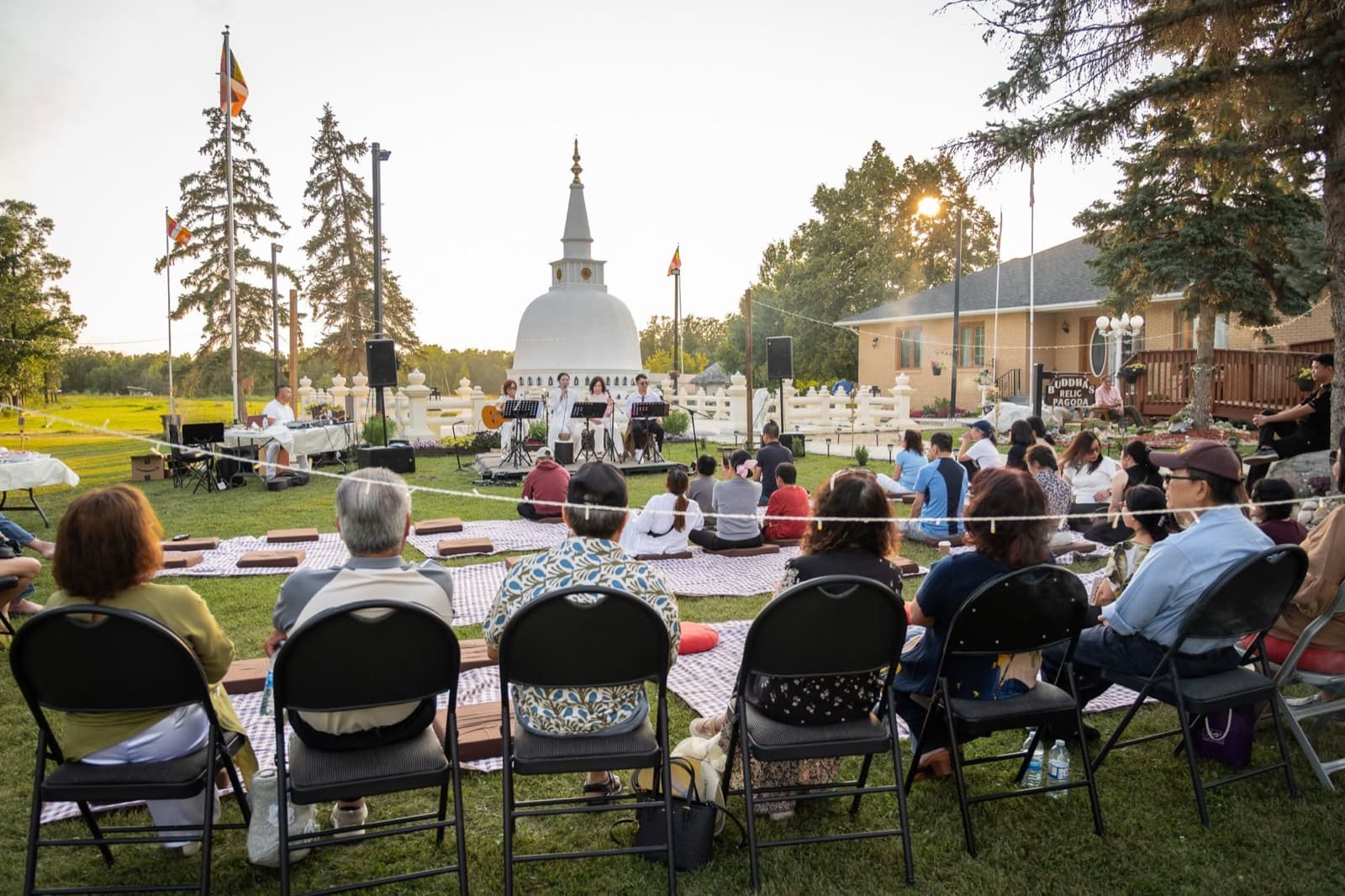 The audience seated on blankets and chairs on the lawn, listening in front of the stupa.