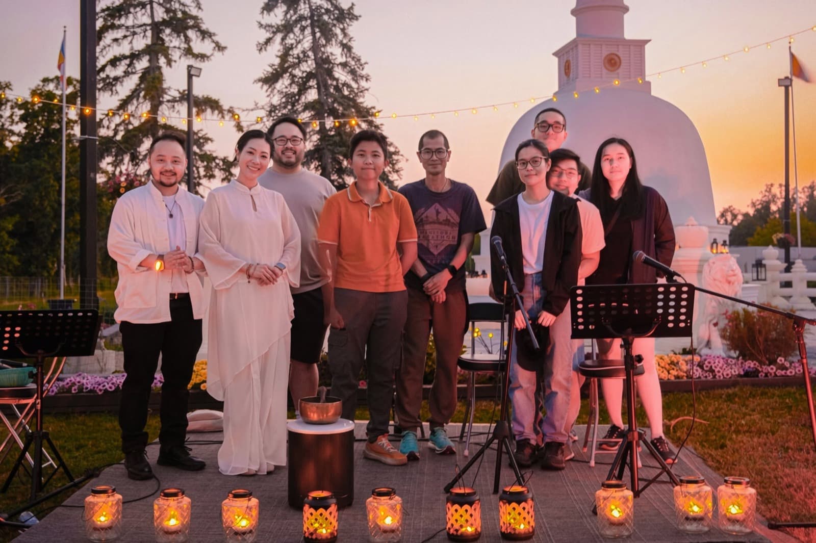The organising team and singers beside a singing bowl under string lights at dusk.